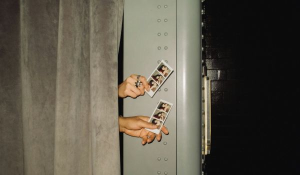 Händer som sticker ut bakom ett sammetsdraperi och håller upp två fotoremsor från en fotoautomat.