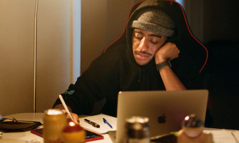 Person i hoodie och mössa skriver i ett block framför en bärbar dator.