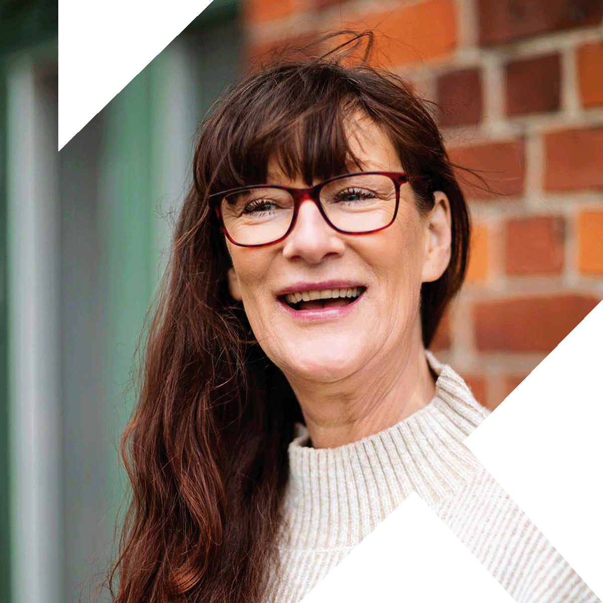 Porträtt av en leende kvinna med långt brunt hår och röda glasögon, bär en beige polotröja. Bilden är inramad av ett stort svart grafiskt K.