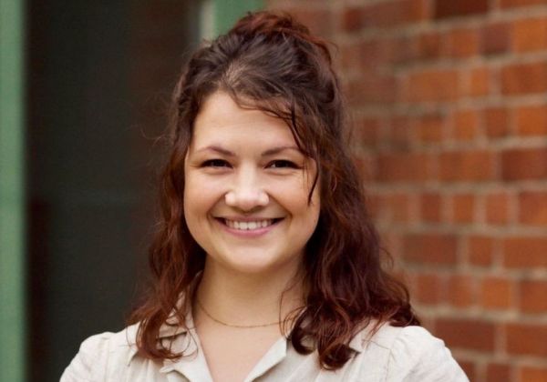 Ett glatt porträtt av Sanna Vuorenmaa med långt, mörkt lockigt hår i en halvuppsättning. Hon bär en ljus blus och står framför en röd tegelvägg.