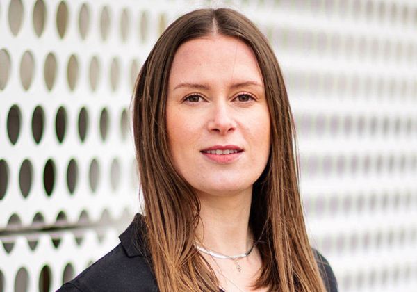 Ett porträtt av Sara Östebro med långt rakt brunt hår och en vänlig blick. Hon bär en svart tröja och ett tunt silverhalsband framför en vit vägg med runda hål.