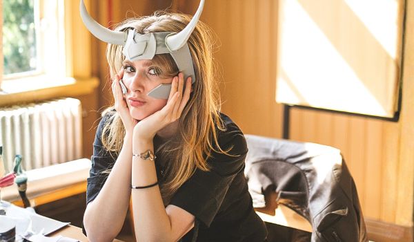 Person med hornförsedd hjälm vilar huvudet i händerna vid ett arbetsbord med pysselmaterial.