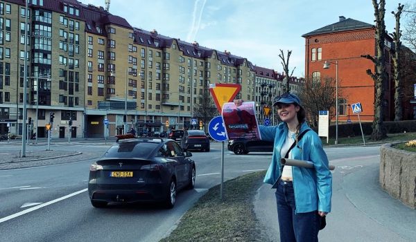 Ung tjej i blå jacka står vid en trafikerad väg och visar stolt upp en affisch med text.