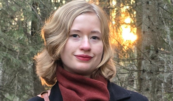 Tilia Thorell med röd halsduk står framför granar med mjukt kvällsljus bakom sig