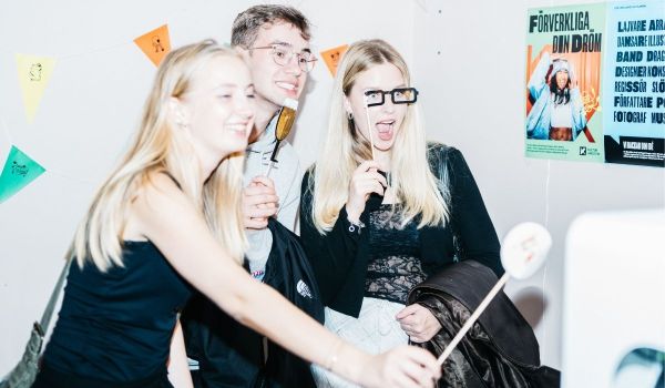 Tre glada ungdomar spexar med fotorekvisita framför en vit vägg med vimplar och affischer.