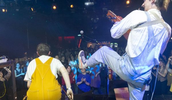 Artister i gula och blå hängselbyxor bjuder på energifylld folkmusik på en scen med publikkontakt.