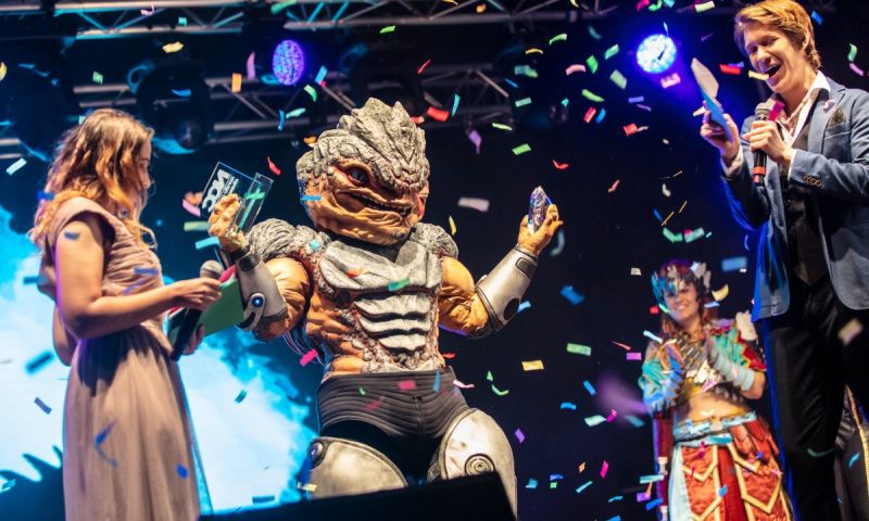 En cosplayer i detaljrik reptilliknande rustning håller upp två priser på en scen medan färgglad konfetti regnar ner och personer runt omkring firar.