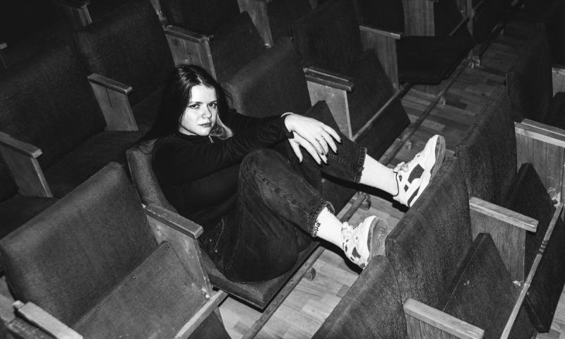 Svartvit bild på en ung kvinna som sitter i en tom biosalong och vilar fötterna på sätet framför.