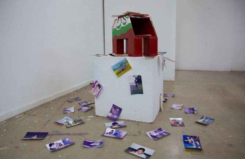 Installation i galleri. En trähäst på en vit låda omgiven av fotografier utspridda på golvet.