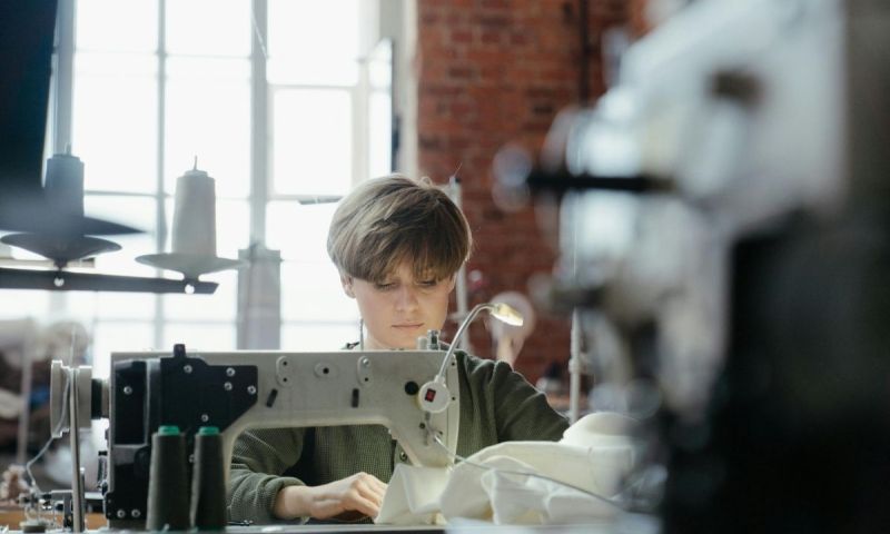 En person syr i vitt tyg med en industrisymaskin i en lokal med tegelväggar.