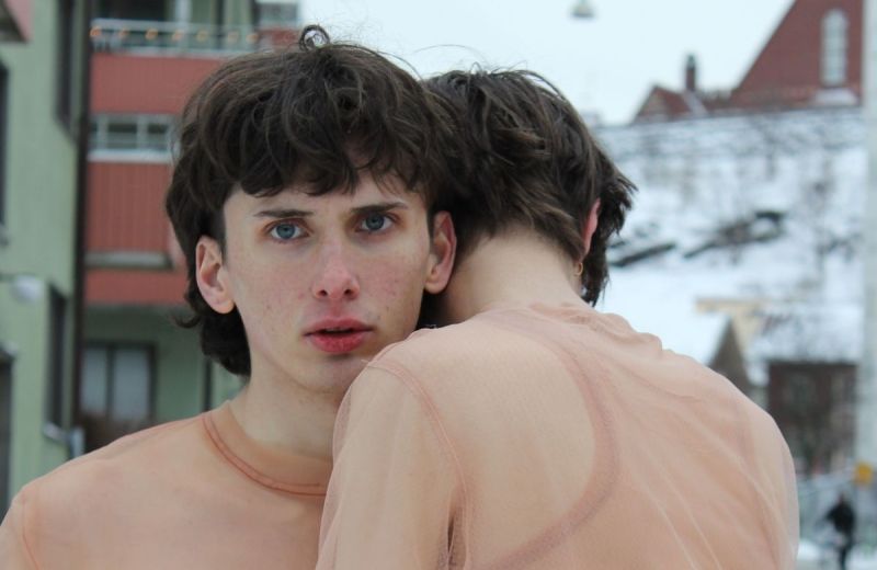 Två personer i hudfärgade nätkläder kramas utomhus i en stadsmiljö.