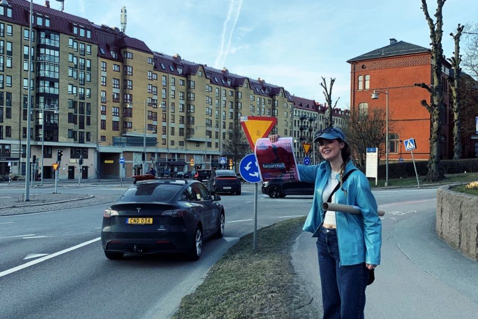 Ung tjej i blå jacka står vid en trafikerad väg och visar stolt upp en affisch med text.