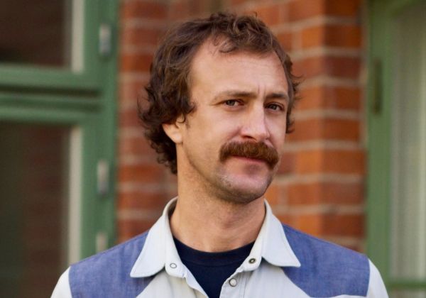 Ett porträtt av Axel Sjöberg med kort brunt lockigt hår och en markant mustasch. Han bär en ljusblå skjorta över en mörk tröja och står framför en grön dörr och tegelvägg.