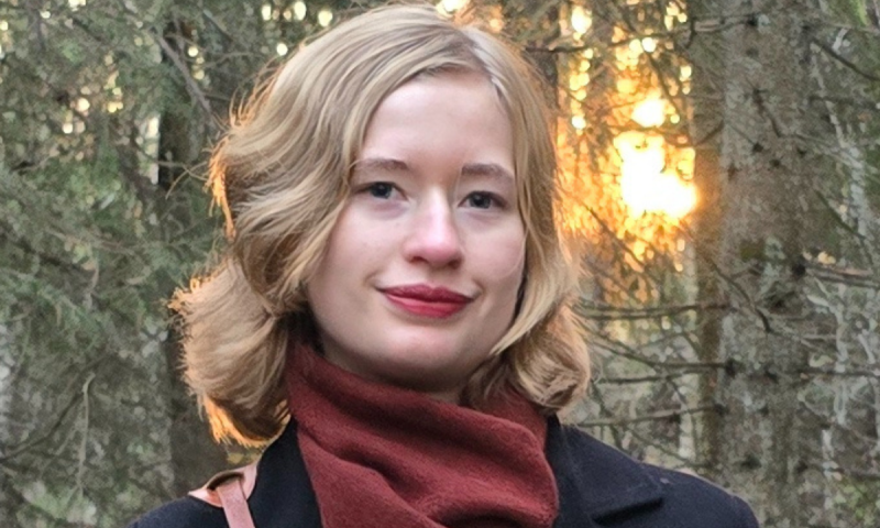 Tilia Thorell med röd halsduk står framför granar med mjukt kvällsljus bakom sig