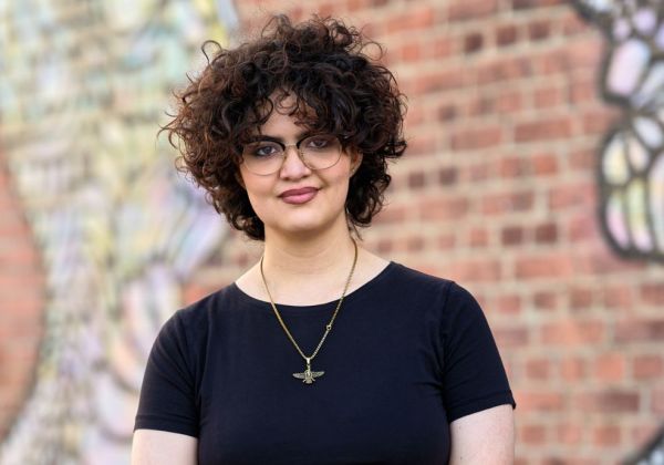 Porträtt av Helen Ashtari med mörkt, kort lockigt hår och glasögon. Hon bär en svart t-shirt och ett guldhalsband framför en graffitimålad tegelvägg.