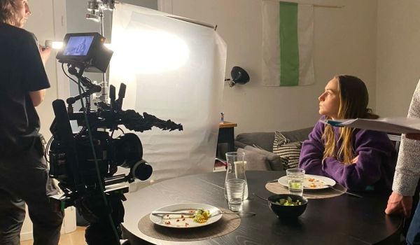 Koncentrerad kvinna i lila tröja vid ett matbord filmas med stor kamera och lampa
