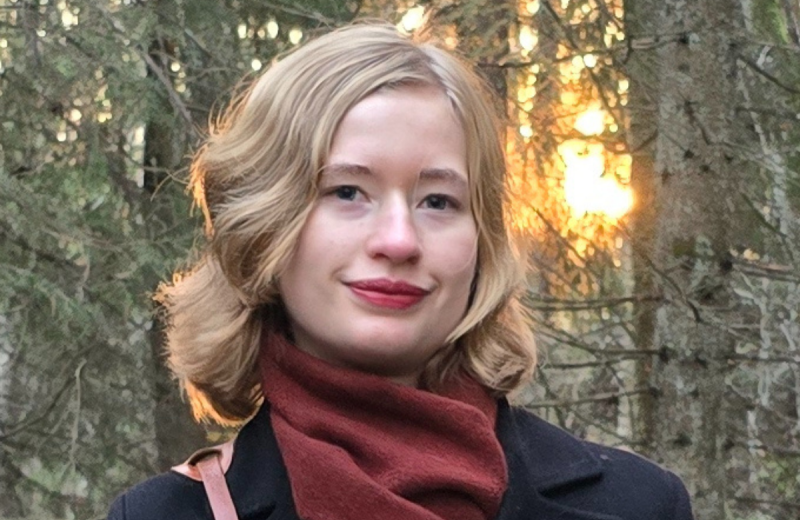 Tilia Thorell med röd halsduk står framför granar med mjukt kvällsljus bakom sig