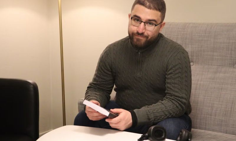 Albaraa Abdulrahman sitter i en soffa vid ett runt vitt bord och håller i sin bok. Han har glasögon, mörkgrön stickad tröja och jeans. På bordet ligger ett par hörlurar.