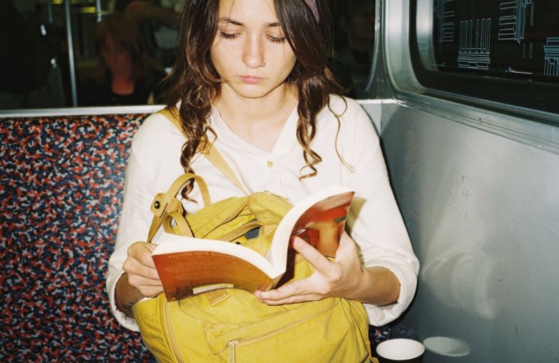 En person sitter på en tunnelbana och läser en bok med en gul ryggsäck i knäet.