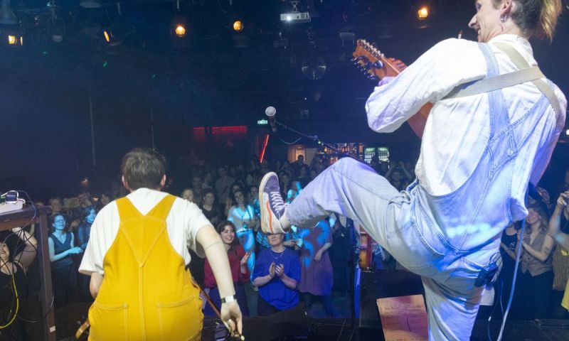 Artister i gula och blå hängselbyxor bjuder på energifylld folkmusik på en scen med publikkontakt.