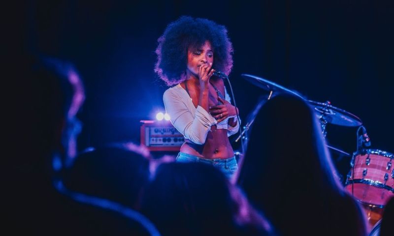 Artist framträder live i ett stämningsfullt scenljus med trummor och förstärkare i bakgrunden.