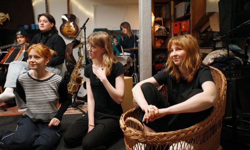 En grupp unga personer i en kreativ repetitionsmiljö med instrument som saxofon, gitarr och dragspel i bakgrunden. En person sitter i en stor flätad korg och ler.