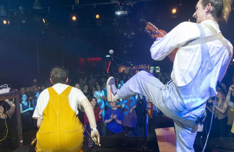 Artister i gula och blå hängselbyxor bjuder på energifylld folkmusik på en scen med publikkontakt.