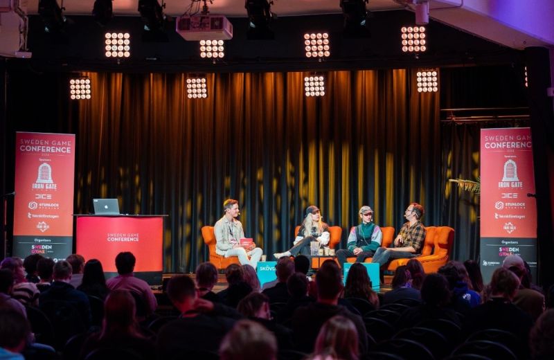 Paneldiskussion under Sweden Game Conference med deltagare i orangea fåtöljer och en stor publik i mörkret.