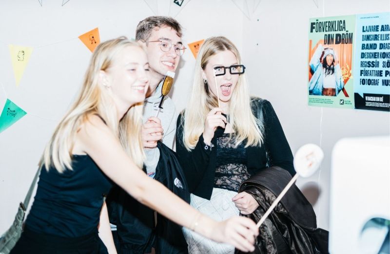 Tre glada ungdomar spexar med fotorekvisita framför en vit vägg med vimplar och affischer.